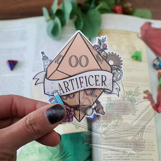 Sticker with 'Artificer' design held by a hand on an open book with decorative elements.