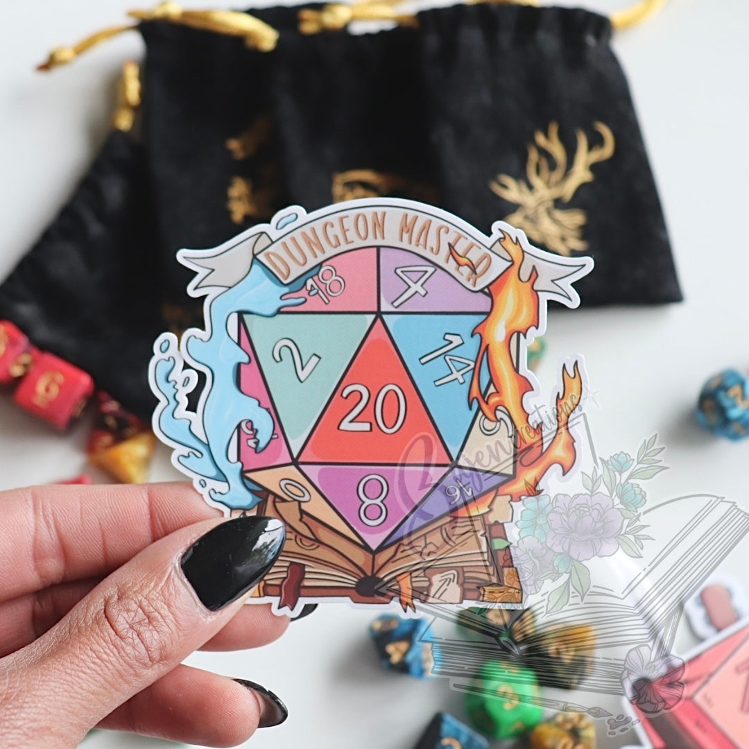Sticker with 'Dungeon Master' text and D20 design held by a hand, with dice and bags in the background.