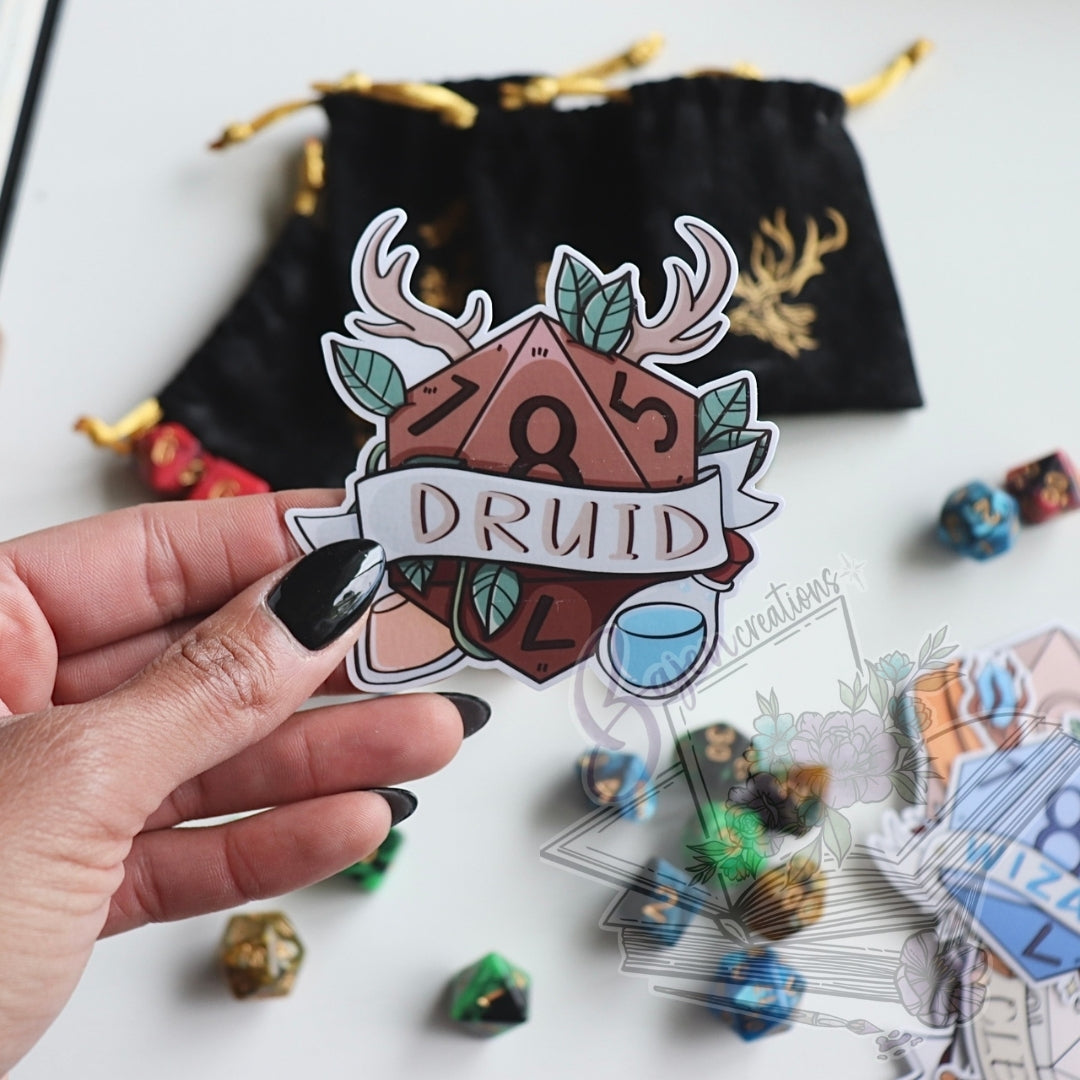 Hand holding a sticker with 'Druid' design, surrounded by dice and cards on a white background
