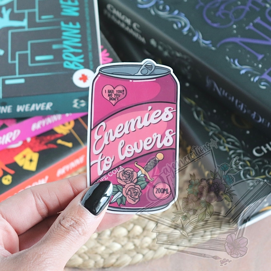 Pink can-shaped product with 'Enemies to Lovers' text held by a hand, with books in the background.