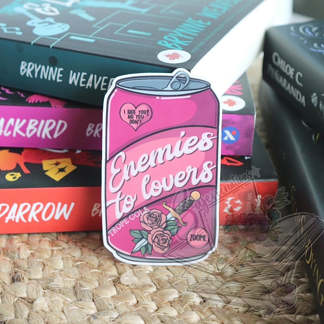 Pink can-shaped product with 'Enemies to Lovers' text, surrounded by books on a textured surface.