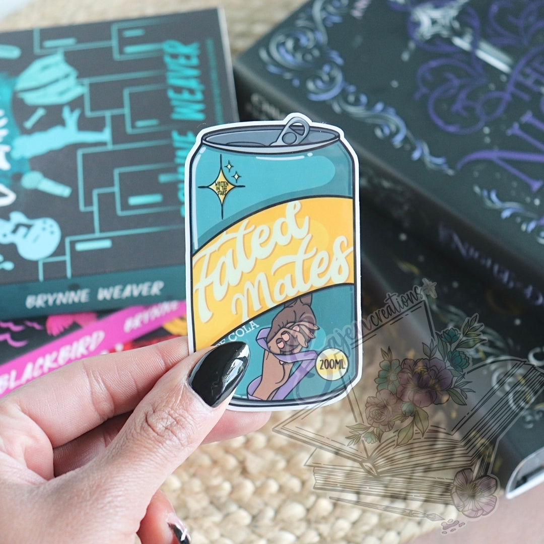 Sticker of a can labeled 'Fated Mates' held by a hand with a book and other items in the background.