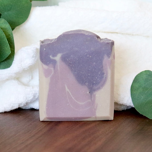 Bar of soap with a purple and white swirl pattern on a wooden surface with green leaves and a white towel in the background.