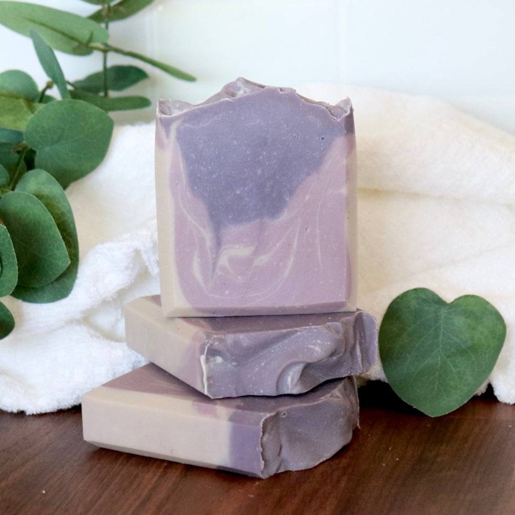 Three bars of purple soap stacked on a wooden surface with green leaves and a white towel in the background.