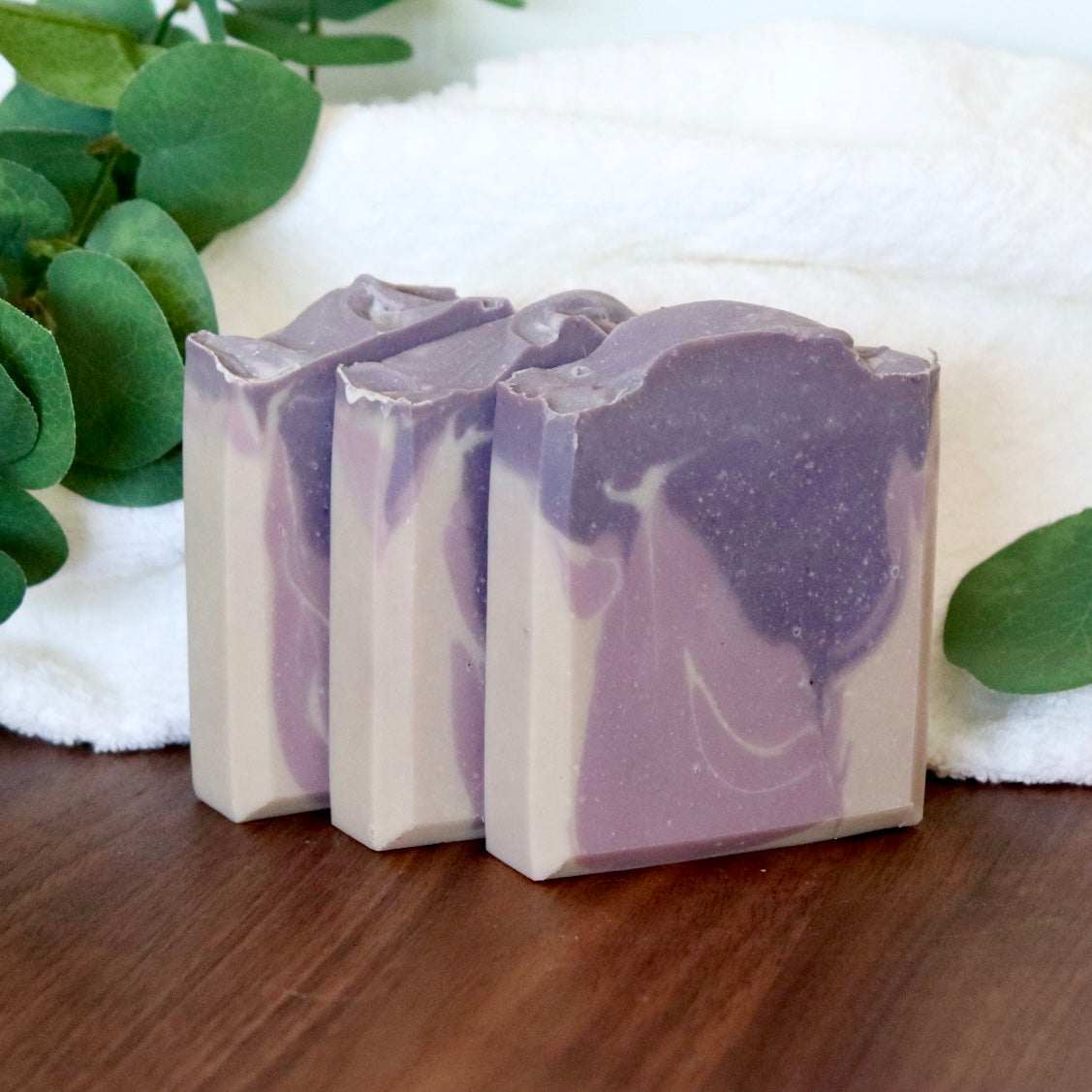 Three bars of purple and white soap on a wooden surface with green leaves in the background
