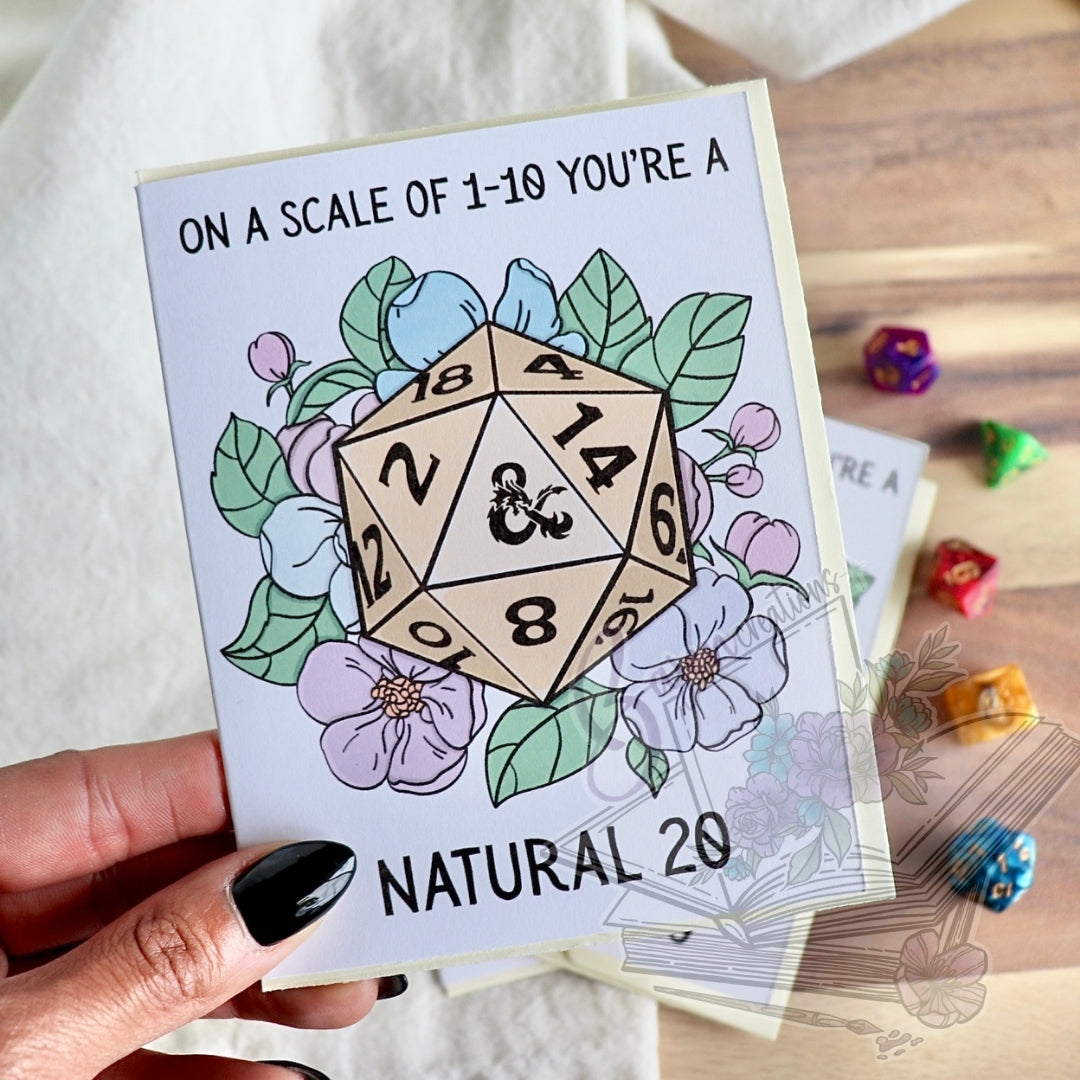 Card with a D20 die illustration and floral elements, held by a hand on a wooden surface with dice.
