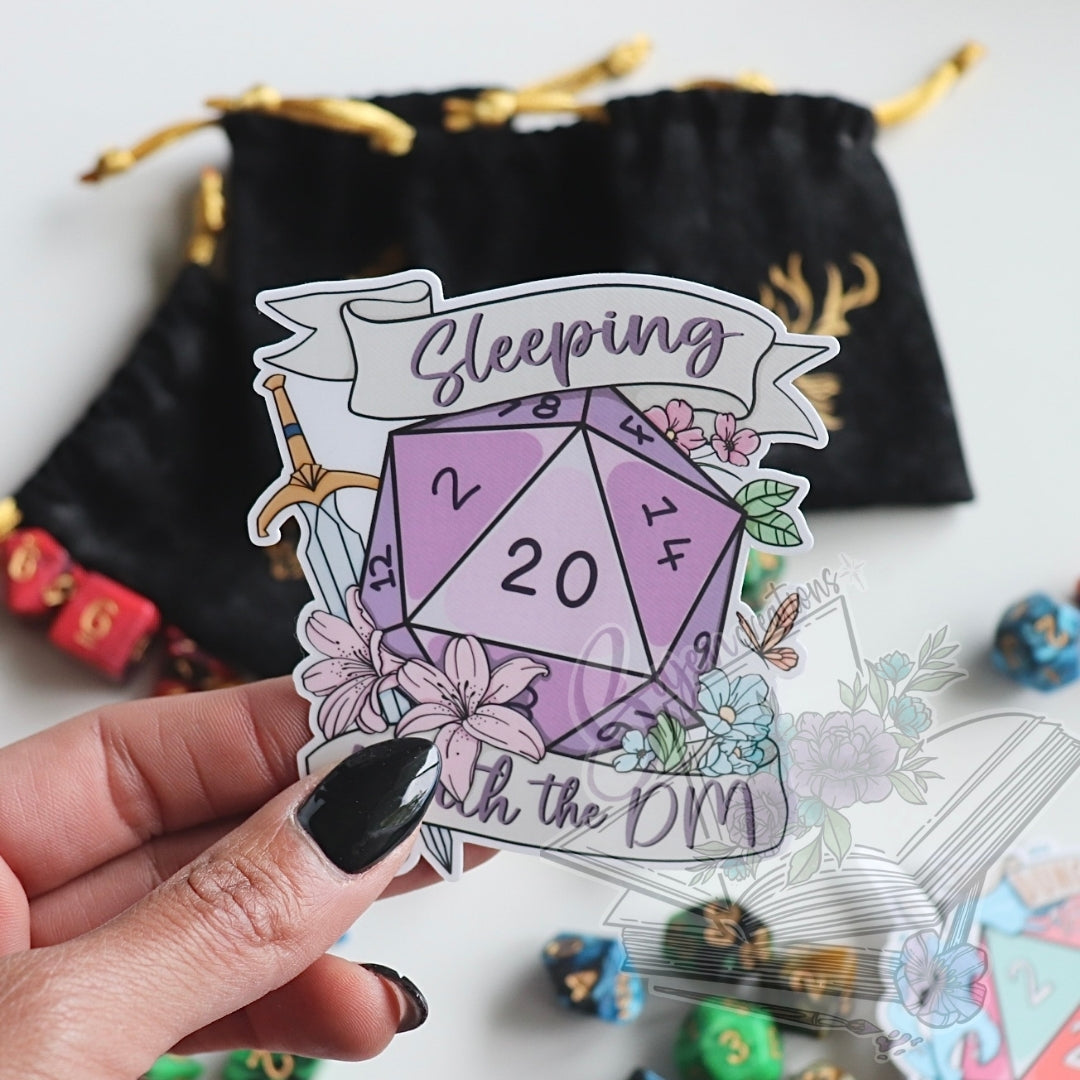 Sticker with a purple die and floral design held by a hand, with dice and a drawstring bag in the background.