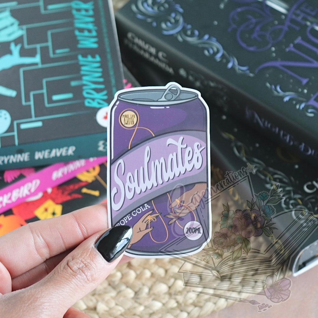 Sticker of a purple can labeled 'Soulmates' held by a hand with black nail polish, against a blurred background of books.