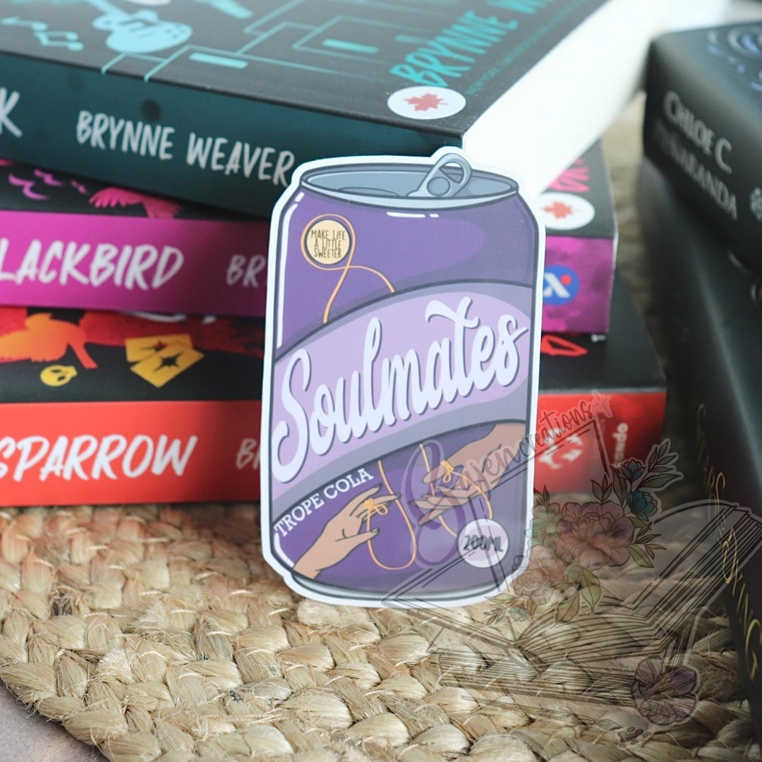Purple can-shaped sticker with 'Soulmates' text on a woven surface with books in the background